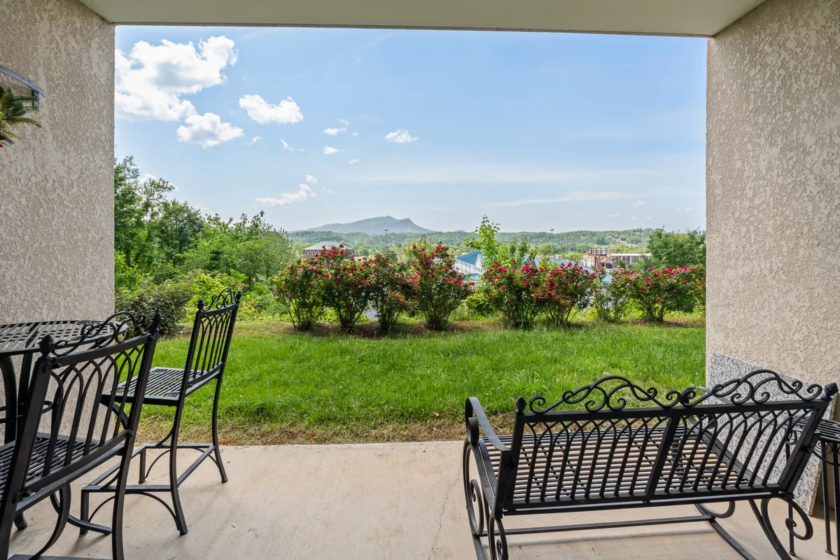 An inviting outdoor space with metal benches placed on lush green grass, surrounded by flowering bushes and framed by a stunning mountain view beneath a blue sky. The setting is perfect for relaxation after exploring nearby attractions.