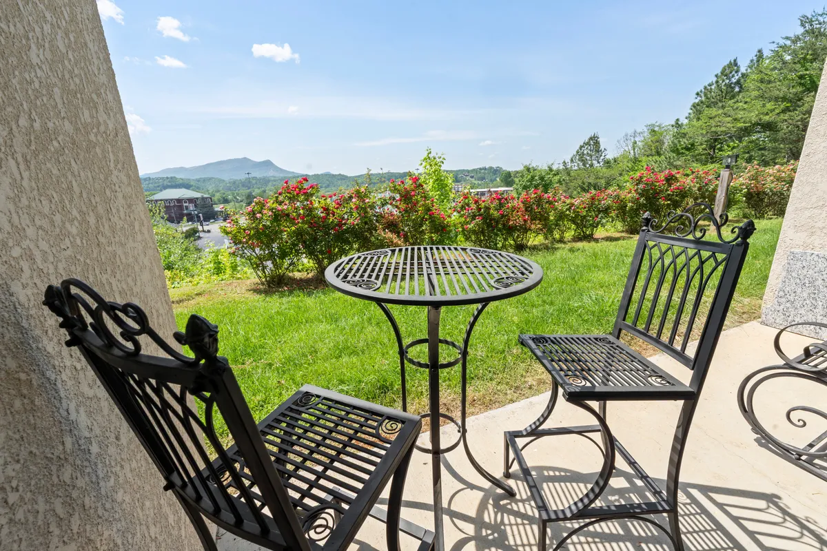 A serene outdoor patio area with a metal table and two chairs framed by flowering bushes, set against a backdrop of breathtaking mountain views. The setting is intimate and ideal for enjoying a peaceful morning coffee or relaxing in the fresh air while taking in the scenery.