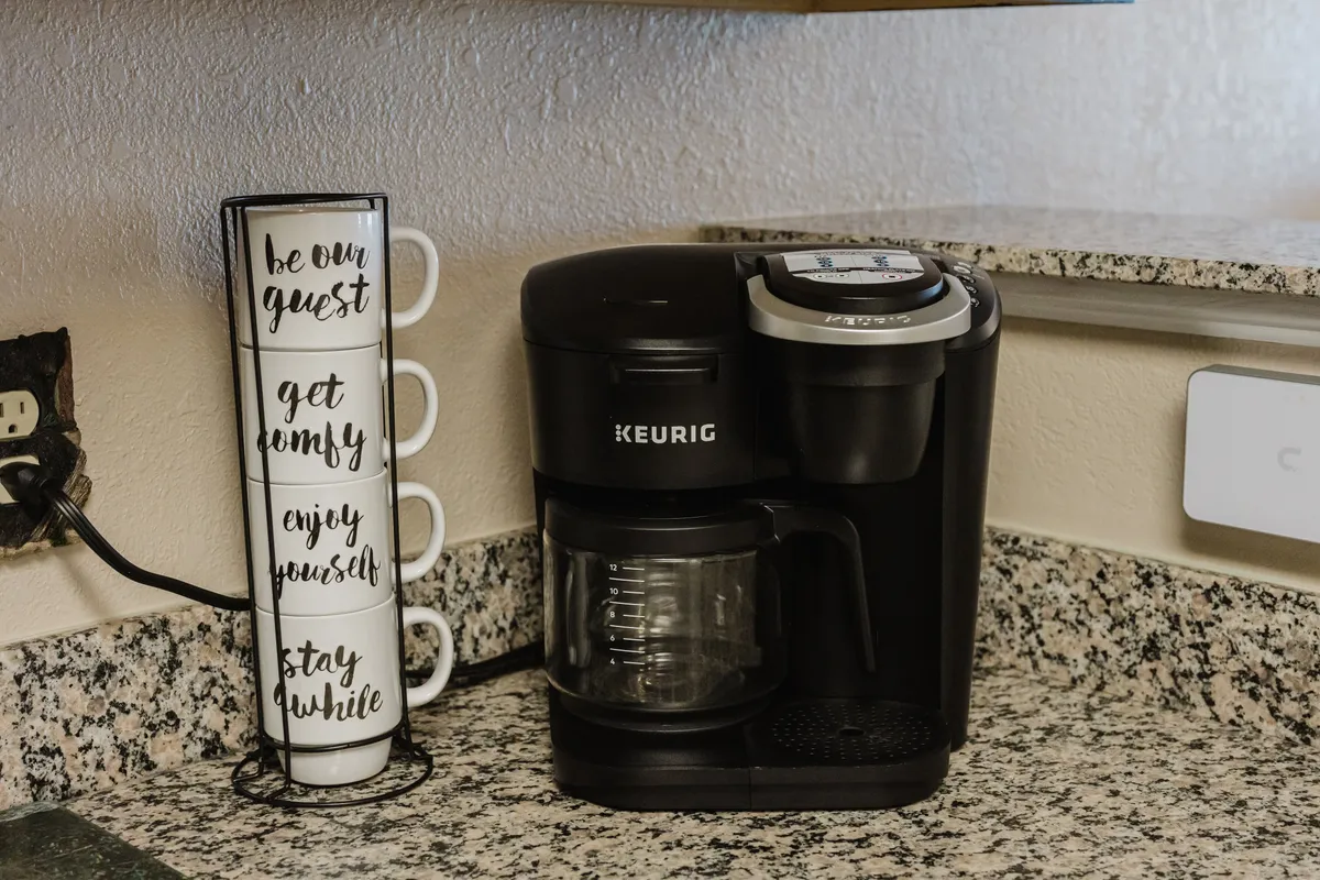 A Keurig coffee maker sits on the kitchen counter beside a stack of inviting cups. The scene feels cozy and welcoming, suggesting easy morning routines and moments of connection over coffee.