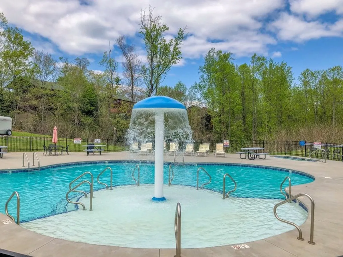 A vibrant outdoor swimming pool scene featuring a central water fountain, several lounge chairs arranged for sunbathing, and lush greenery encircling the pool. The atmosphere is playful and refreshing, ideal for families to gather and enjoy sunny afternoons.