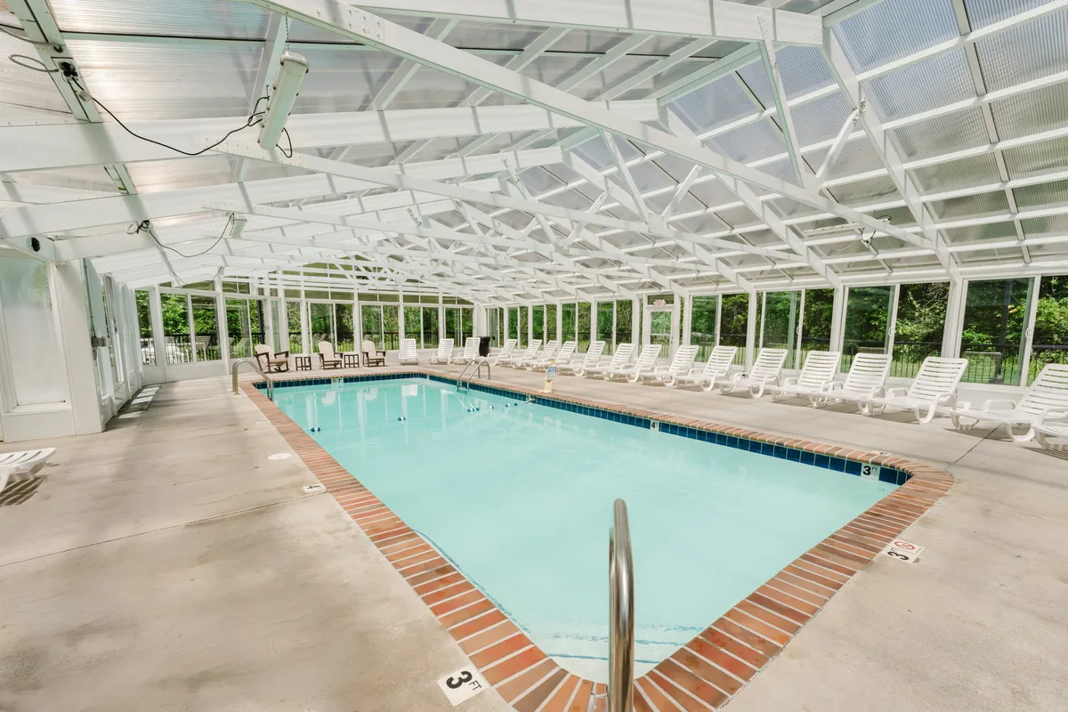 A bright, inviting indoor swimming pool surrounded by lounge chairs and illuminated by a glass ceiling. The space feels airy and relaxed, suggesting year-round swimming and socializing regardless of the weather.