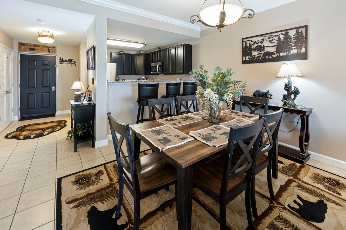 A spacious dining area with a large table set on a decorative rug, bar stools at the counter, and tasteful wall art. The layout is open, allowing for easy flow between dining and adjacent living spaces, perfectly suited for group gatherings or family meals.
