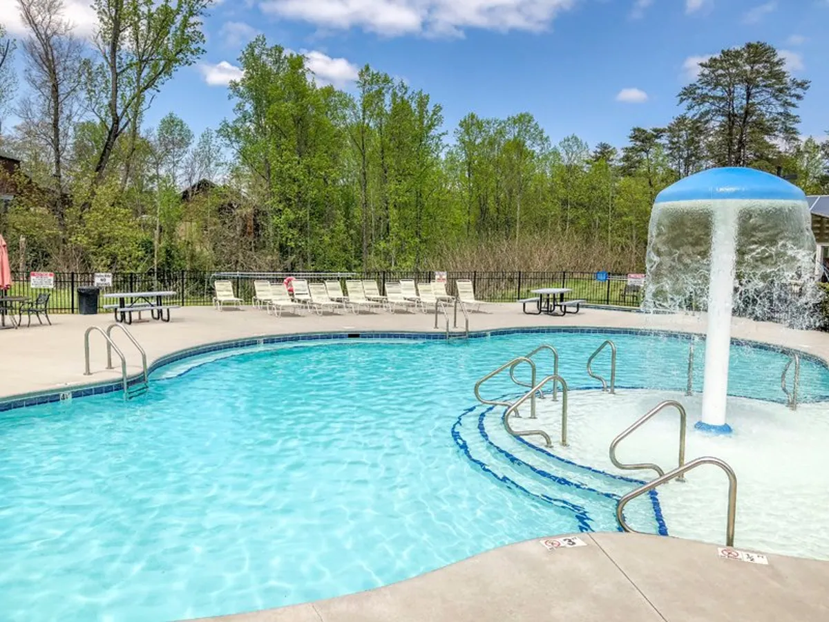 A sparkling outdoor swimming pool with a water feature, surrounded by lush greenery and inviting lounge chairs. The scene exudes leisure, with room for sunbathing, swimming, and social gatherings against a beautiful natural backdrop.