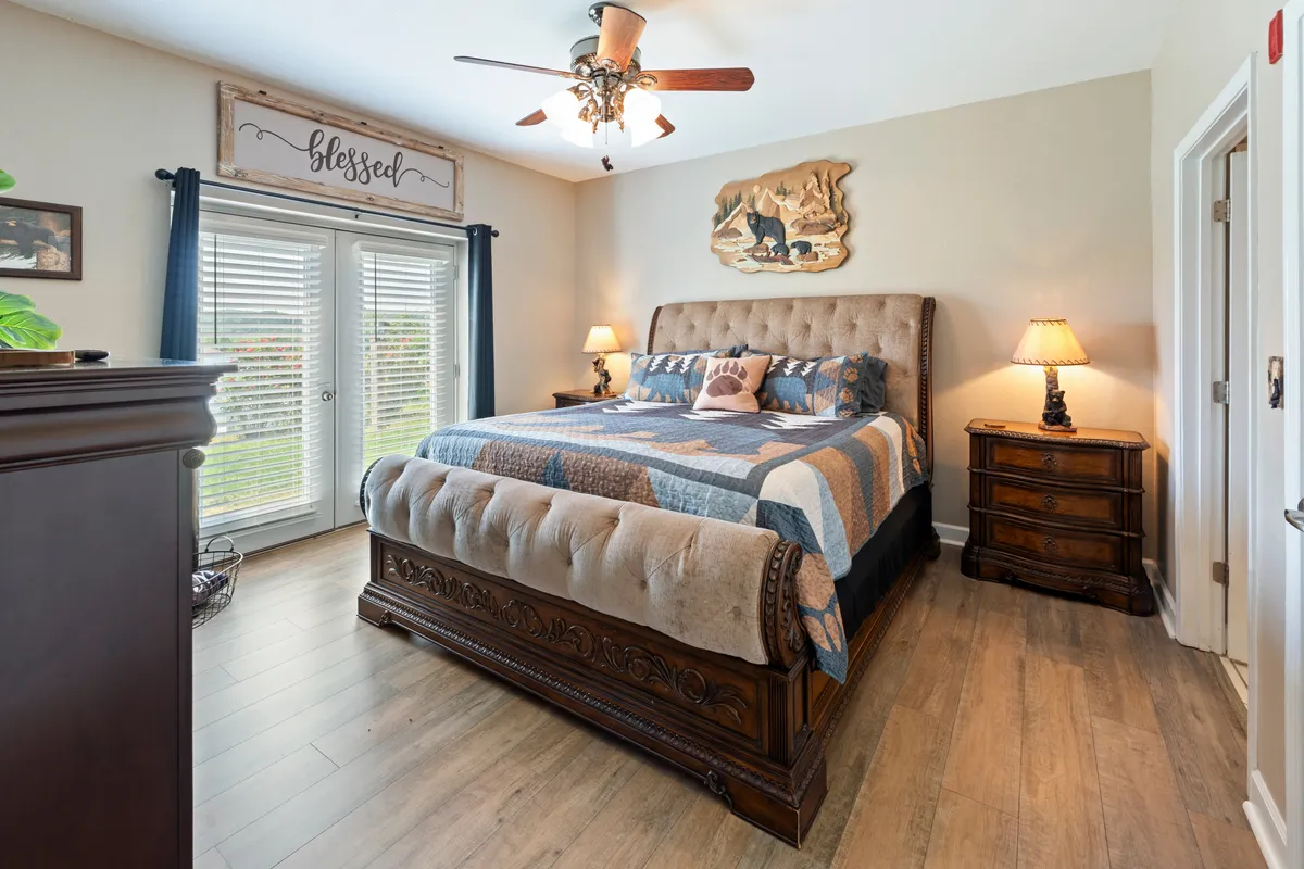 A spacious master bedroom featuring a king-size bed with decorative bedding, wooden bear wall art above the headboard, nightstands with softly glowing lamps on both sides, and sliding glass doors with curtains that offer direct access to an outdoor patio.