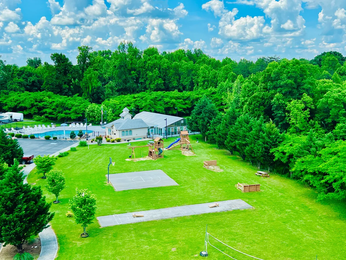 A lively exterior scene featuring playground equipment, a basketball court, a swimming pool, and the main building. Children play, families gather, and guests enjoy multiple amenities in a vibrant, active resort atmosphere.