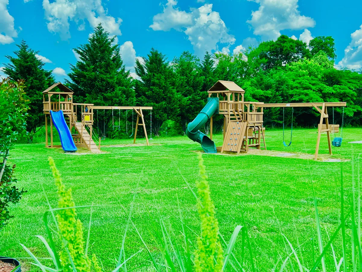 A joyful playground scene showing a colorful playground structure with slides and a swing set set on green grass, with trees in the background. Children are playing, evoking feelings of adventure and playful energy.